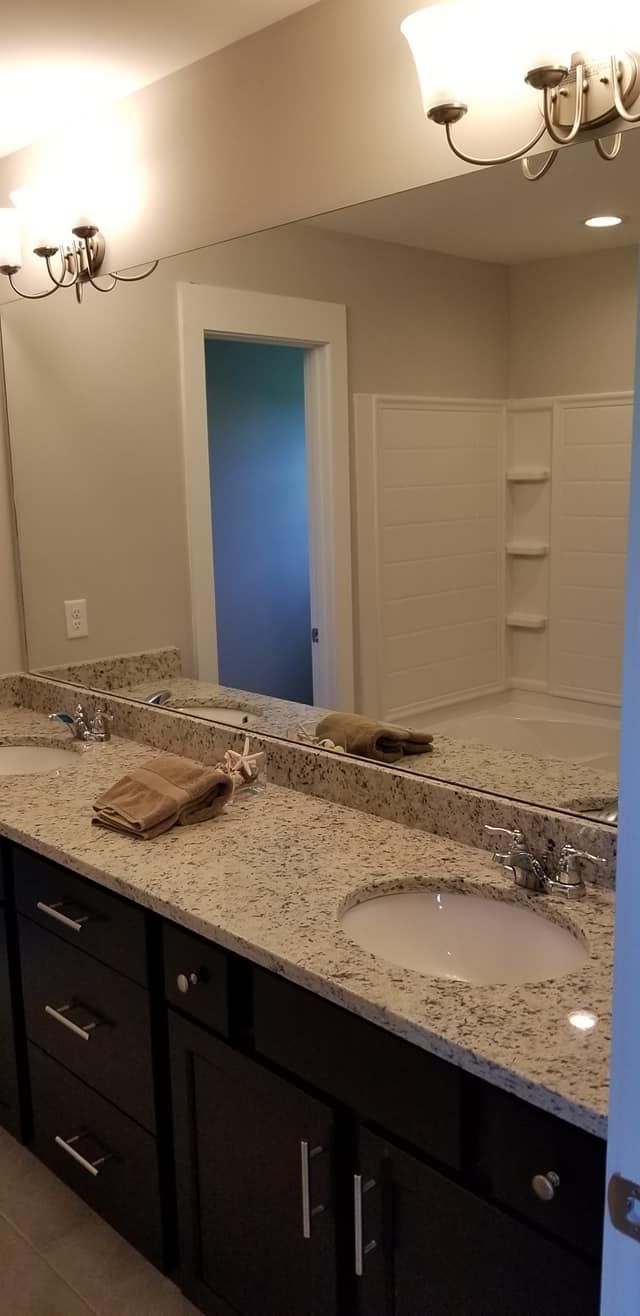 A double-sink bathroom vanity with dark cabinetry and speckled granite countertops, reflecting a shower in a wall mirror.