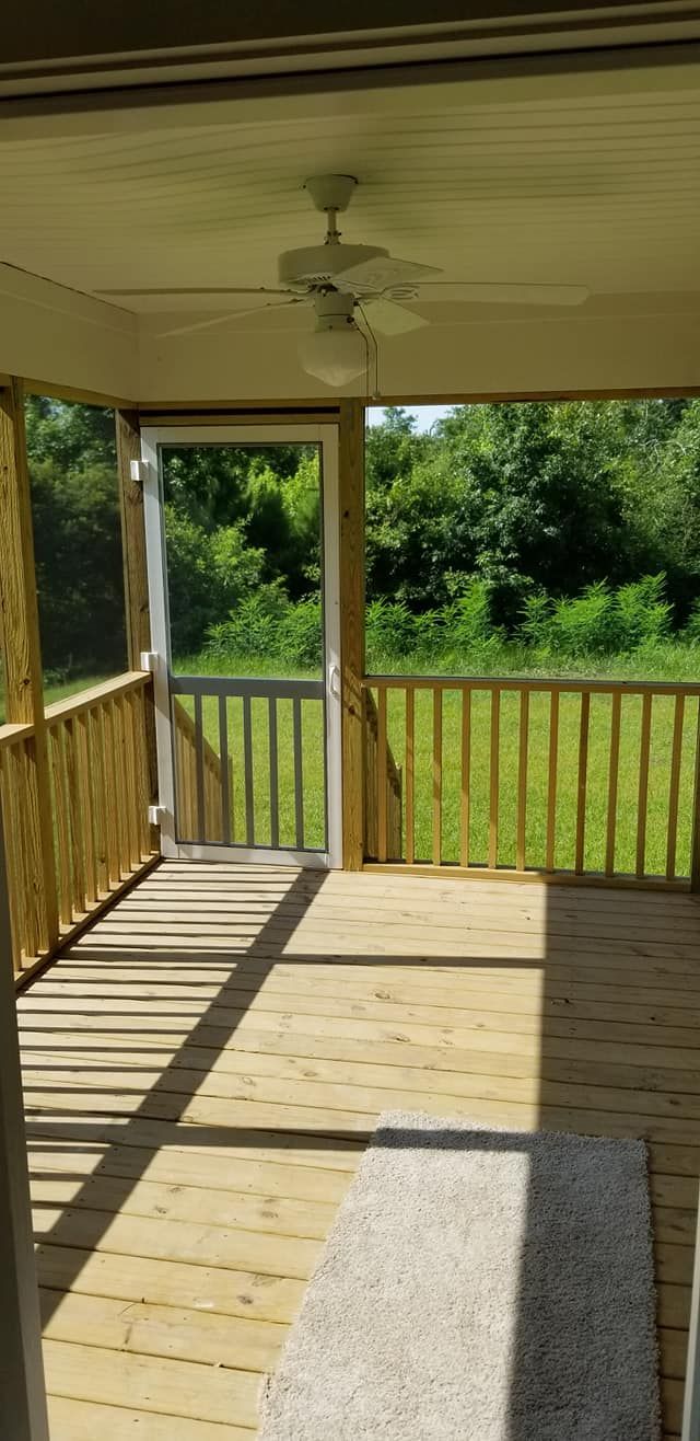 A sunny, screened-in wooden deck with a ceiling fan, a screen door, and a view of trees and grass.