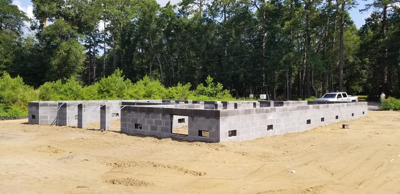 A gray concrete block foundation for a building sits on a sandy construction site in front of a line of green trees.
