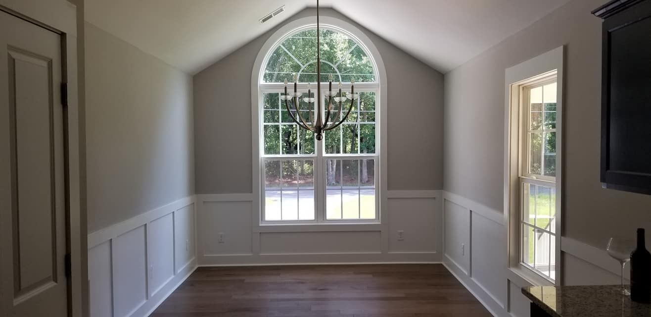 A dining room with white wainscoting, light gray walls, dark wood floors, a large arched window, and a chandelier.