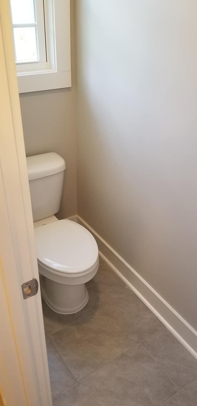 A white toilet in a small, neutral-toned bathroom with light grey flooring and a window on the side wall.