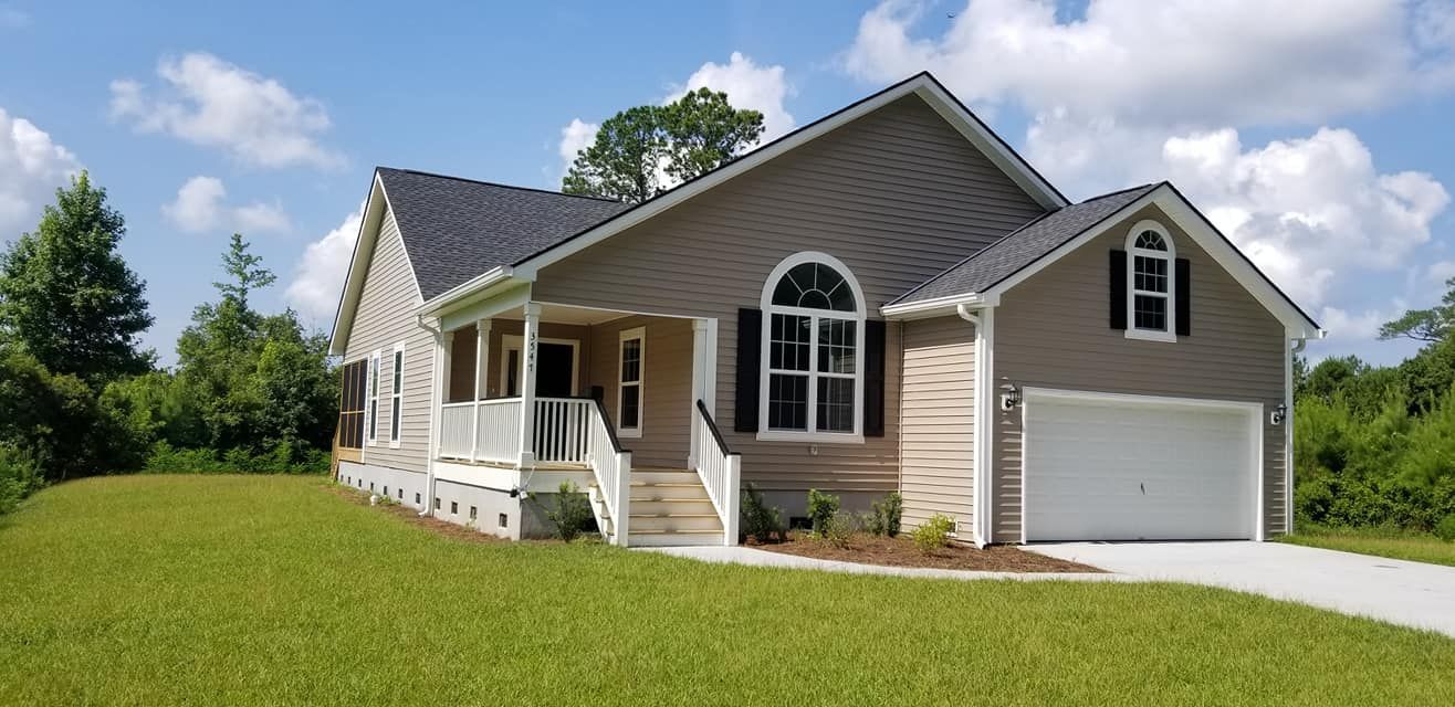 A tan, single-story suburban house with a dark gabled roof, white garage door, and front porch, set in a grassy landscape.