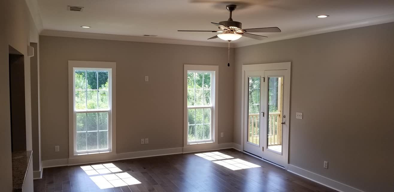 An empty, neutral-toned room with hardwood floors, a ceiling fan, two windows, and glass double doors leading outside.
