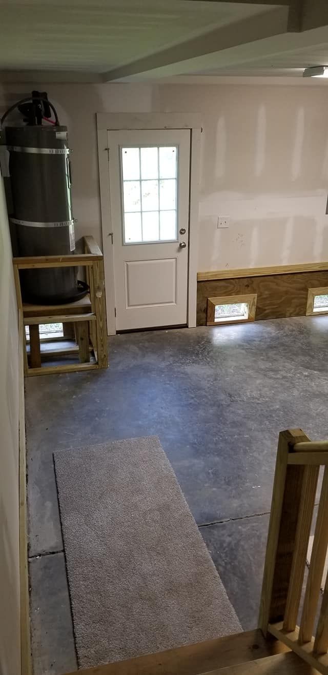 A basement with a concrete floor, a white door, an unfinished wall with exposed insulation, and a water heater on a stand.