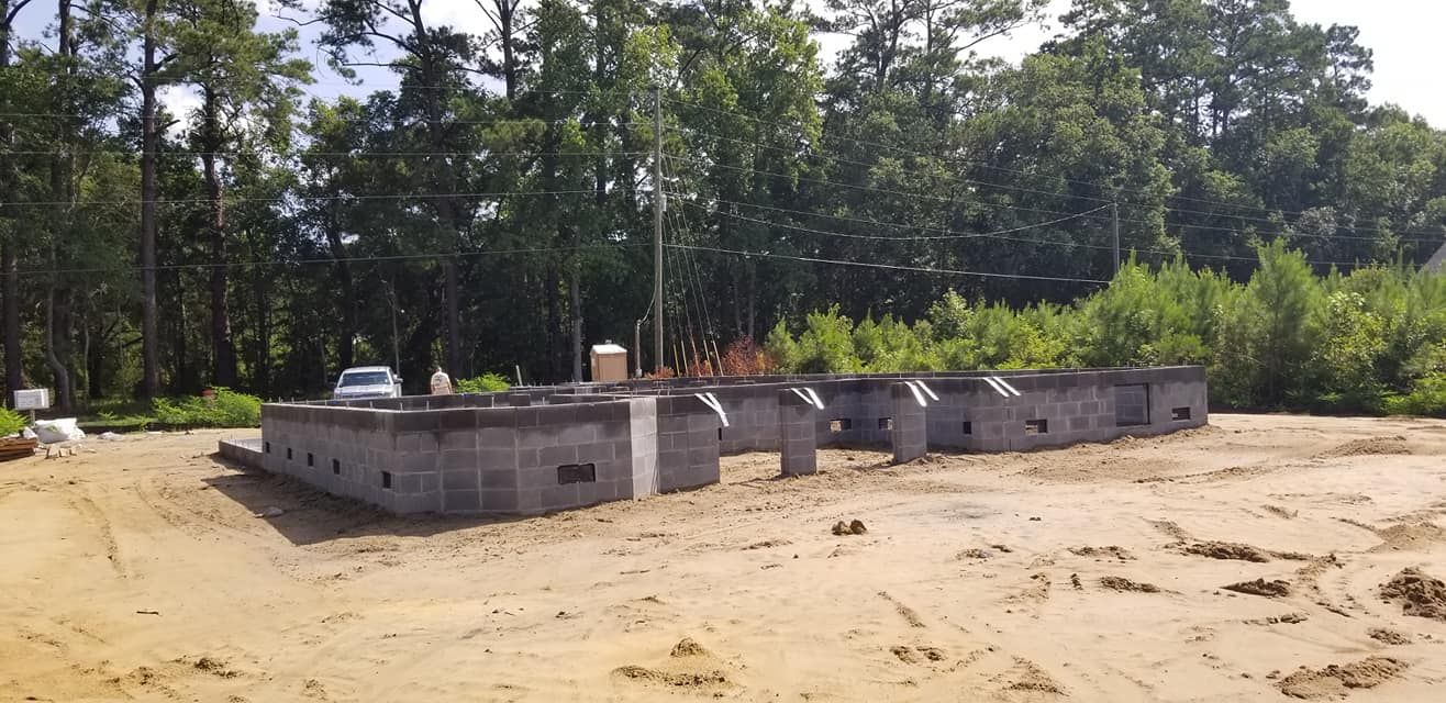 A low-lying concrete block foundation for a building under construction on a sandy lot surrounded by trees.