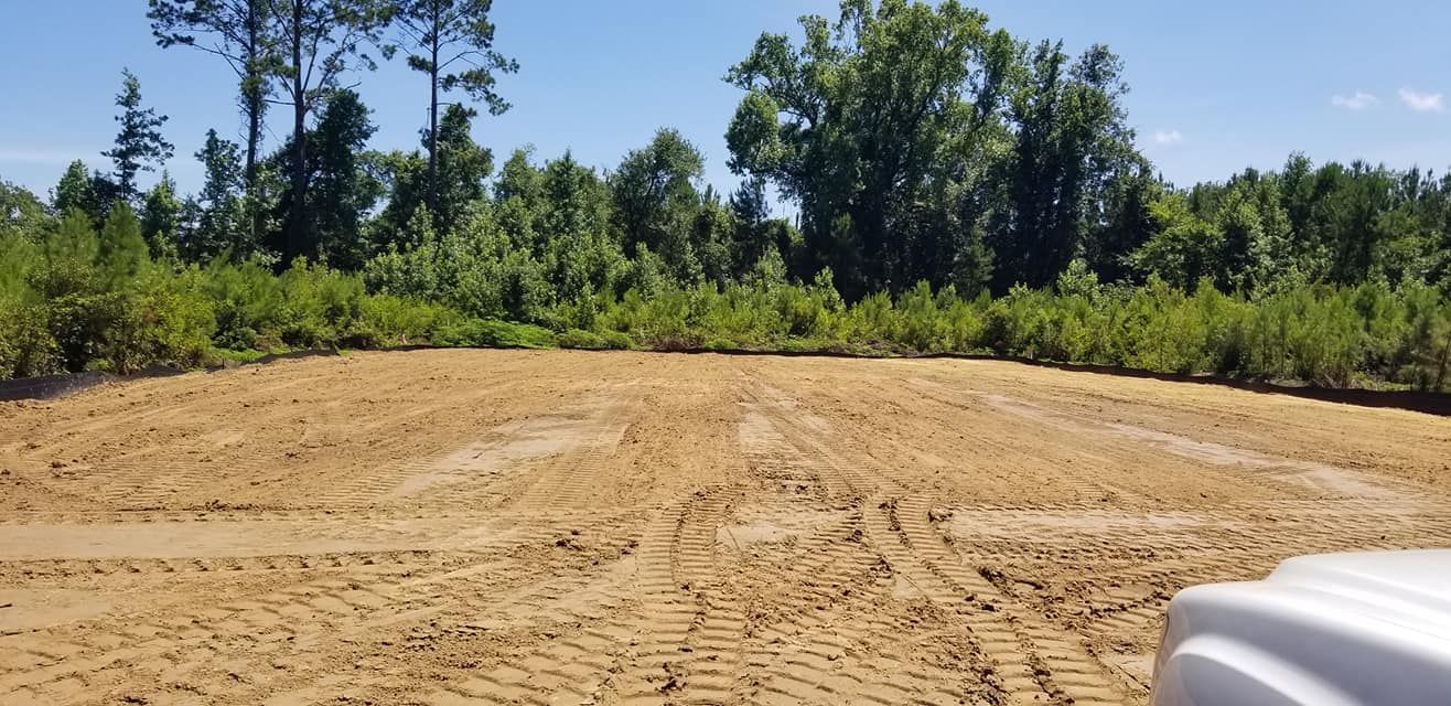 A freshly cleared, sandy lot with tire tracks, bordered by dense green trees under a clear blue sky.