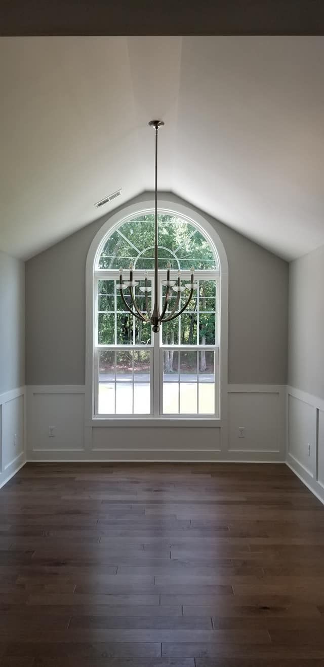 A bright room with wood floors, white wainscoting, light gray walls, a large arched window, and a chandelier.