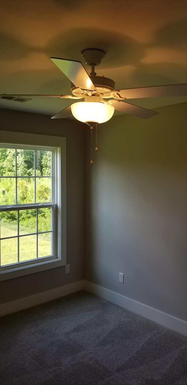 A corner of a room with gray carpet, a white-framed window looking out to greenery, and a lit ceiling fan overhead.