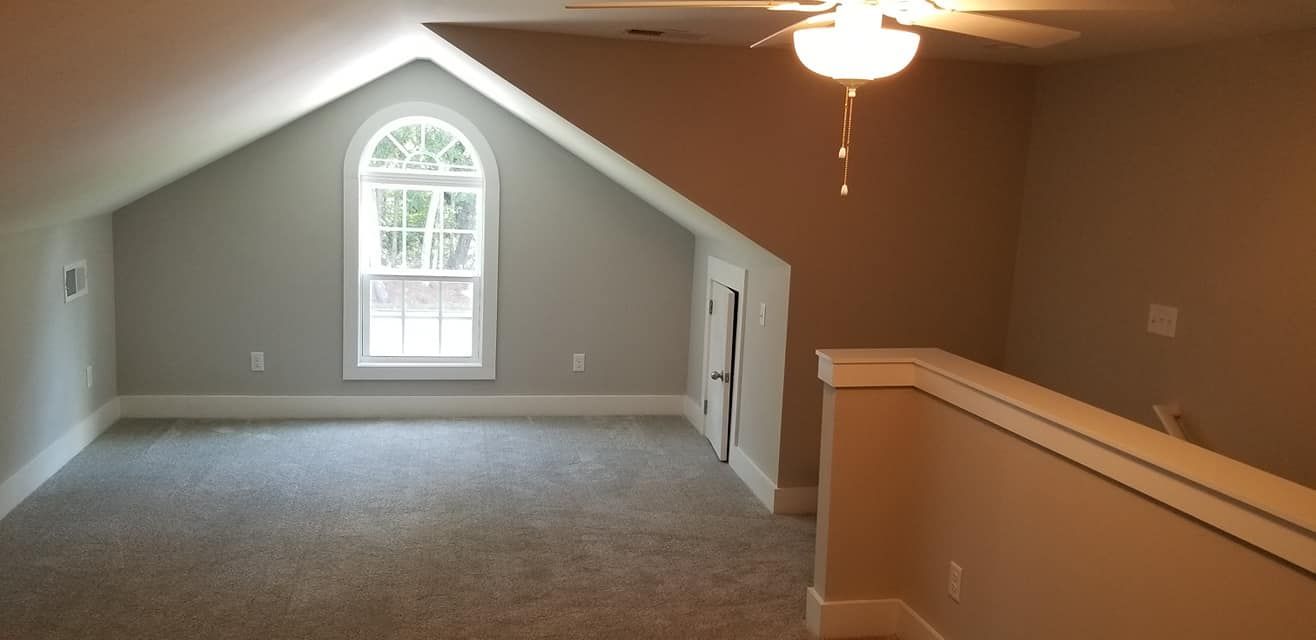 An attic room with a sloped ceiling, gray carpet, a central window, a ceiling fan, and a partial staircase wall.