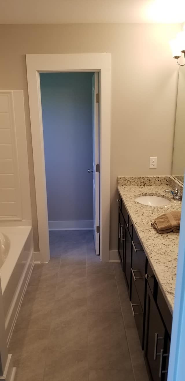 A bathroom with grey tile floors, dark cabinets, granite countertops, and a doorway leading to a small adjacent room.