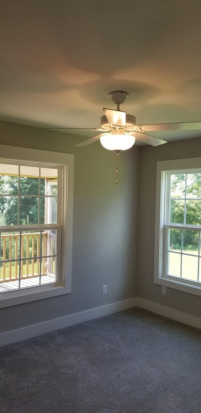 An empty room with gray carpet, gray walls, two windows looking outside, and a white ceiling fan with a light fixture.