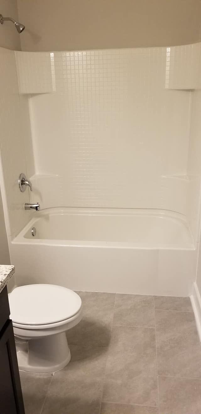 A white bathtub with a showerhead and faucet, next to a white toilet in a bathroom with neutral-toned tiled flooring.