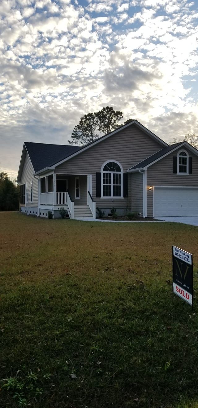 A beige, single-story house with a white garage and front porch stands on a grassy lawn under a cloudy sky.