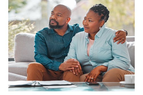 Two people sitting on a couch, holding hands and talking in a bright living room.