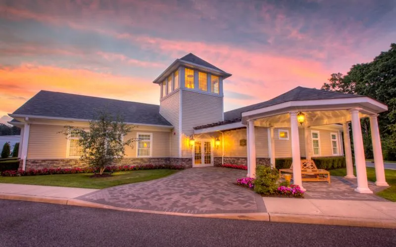 Luxury house at dusk with glowing lights, stone driveway, and pink flowers under a colorful sky