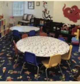 Childcare classroom with round tables, small chairs, blue carpet, and wall decorations.