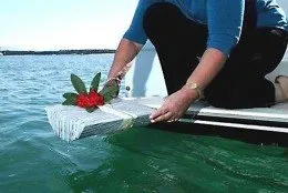 Person kneels at a boat’s edge, placing a white wreath with red flowers into the water