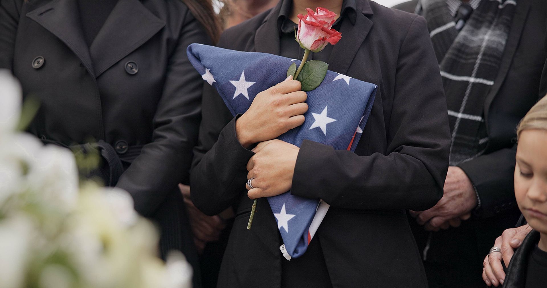 Mourners in dark clothing hold folded U.S. flags and a red rose at a memorial service