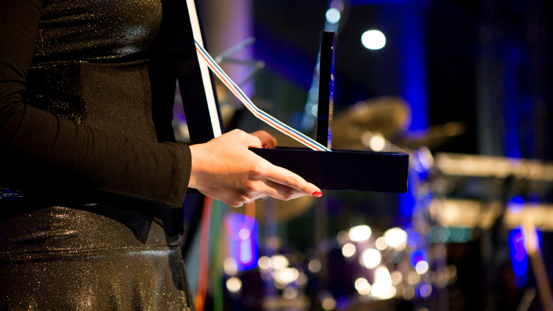 Woman holding an award onstage; black dress, sparkly fabric; band in background.