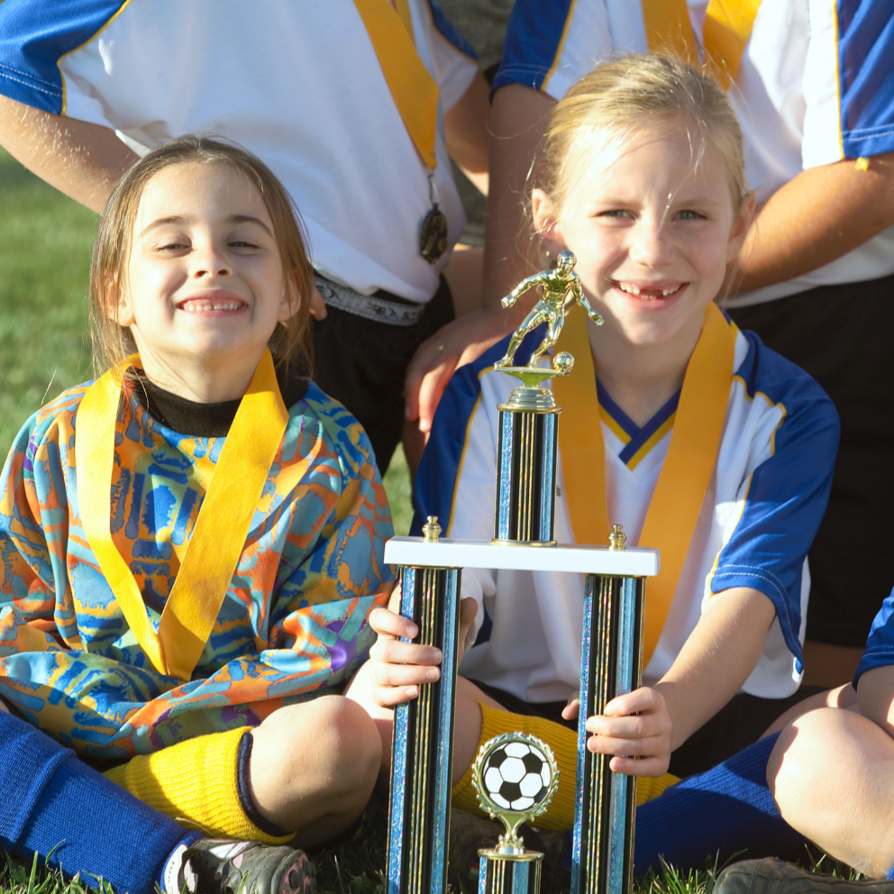 Two girls smiling with a trophy and medals; soccer team on a field.