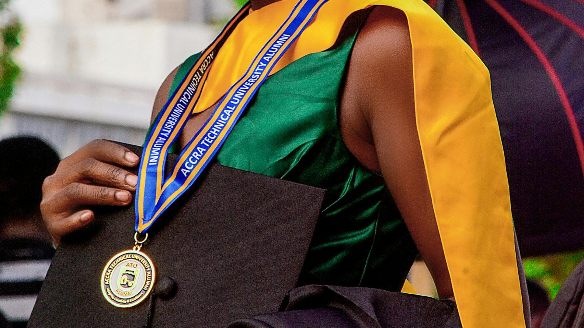 Person in graduation attire, wearing a gold medal with a blue and yellow ribbon. Holding a cap.