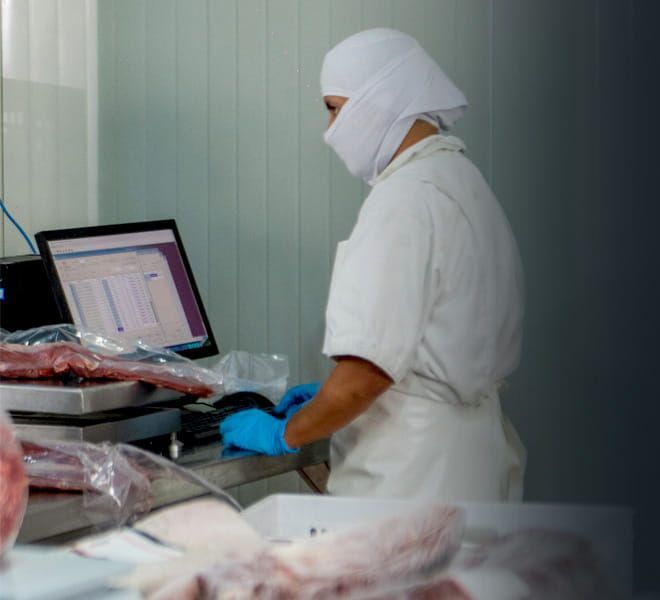 Un trabajador con ropa protectora blanca y guantes clasifica la carne en una mesa de procesamiento junto a la pantalla de un ordenador.