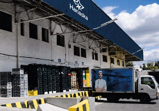 Muelle de carga de un almacén con cajas apiladas y un camión de reparto estacionado junto a un toldo azul.