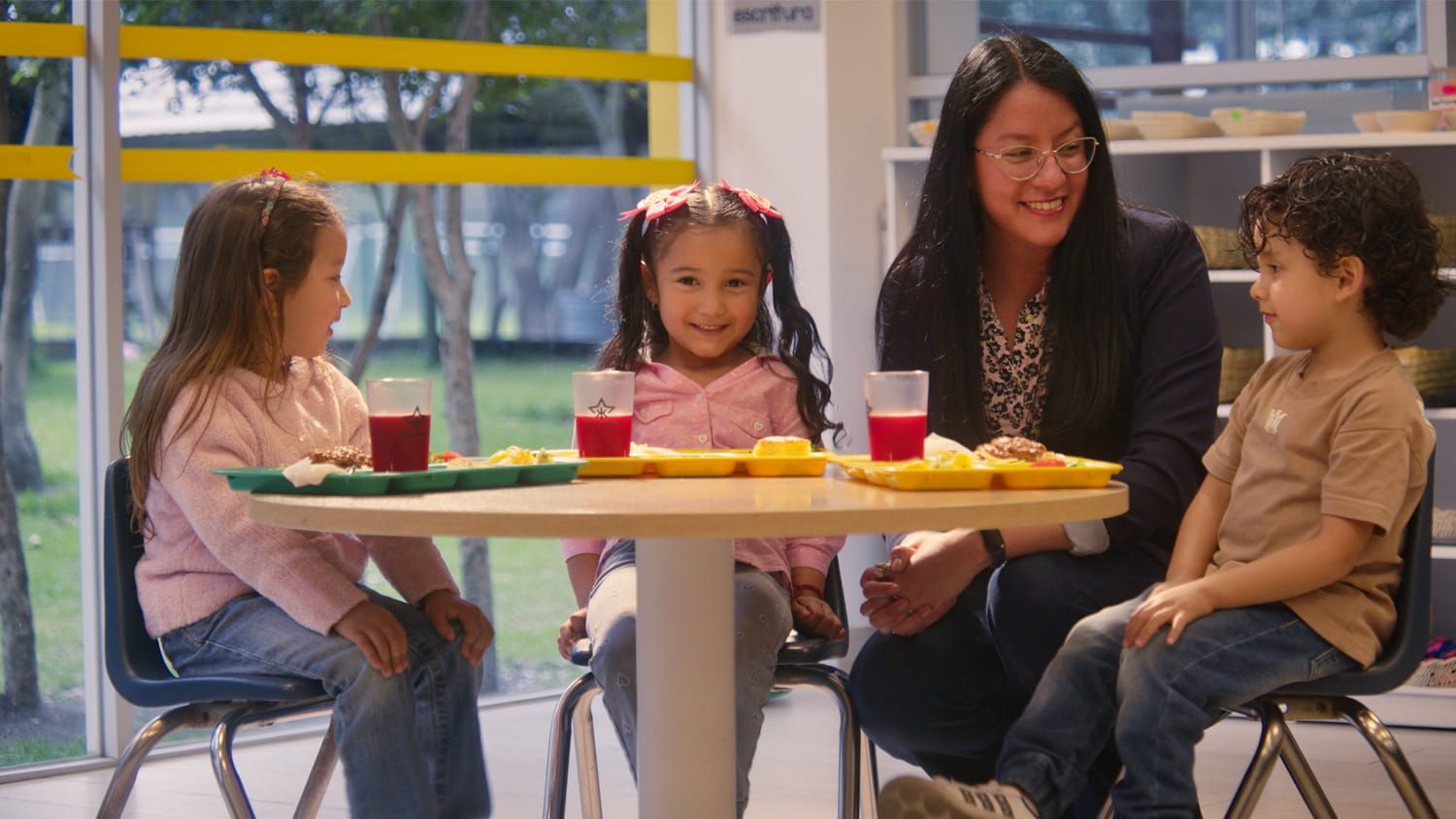 Tres niños están sentados alrededor de una mesa amarilla con bebidas en una luminosa cafetería interior.