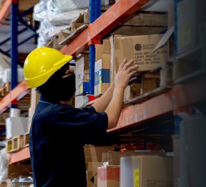 Un trabajador de almacén con casco amarillo intenta alcanzar una caja en un estante de almacenamiento.