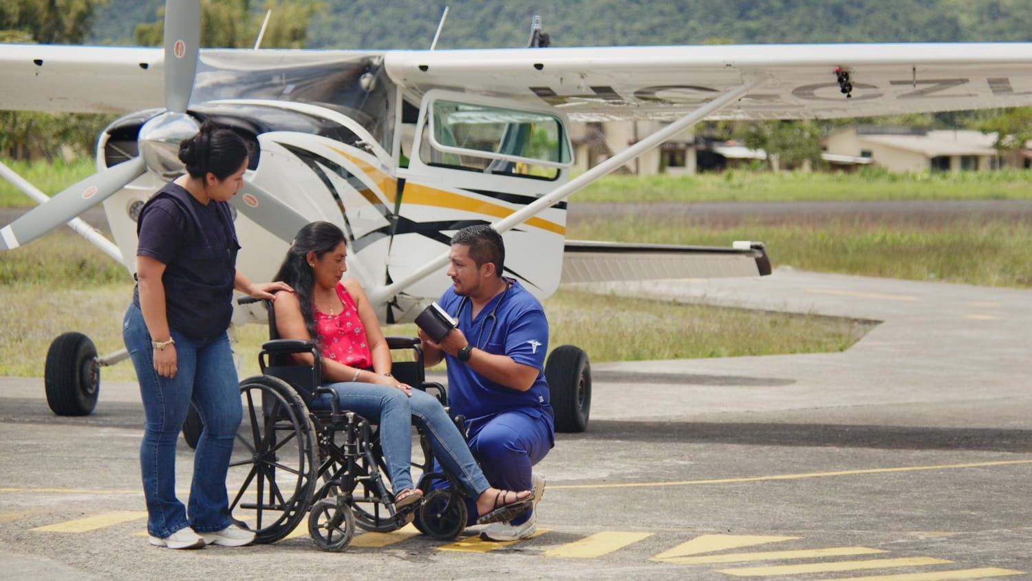 Un hombre con uniforme azul ayuda a una mujer en silla de ruedas junto a una avioneta en la pista de aterrizaje.