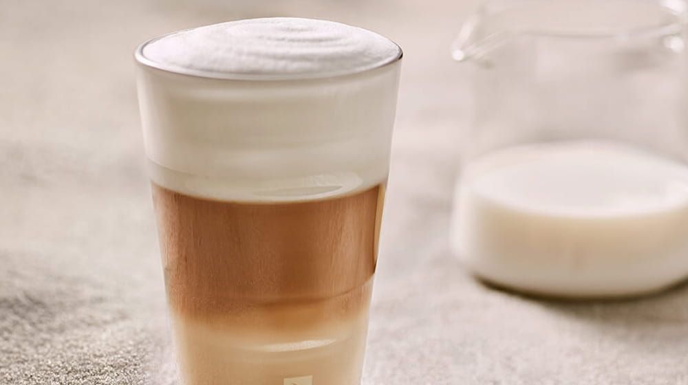 Latte en capas en un vaso con espuma en la superficie, junto a un pequeño plato blanco sobre una superficie beige.
