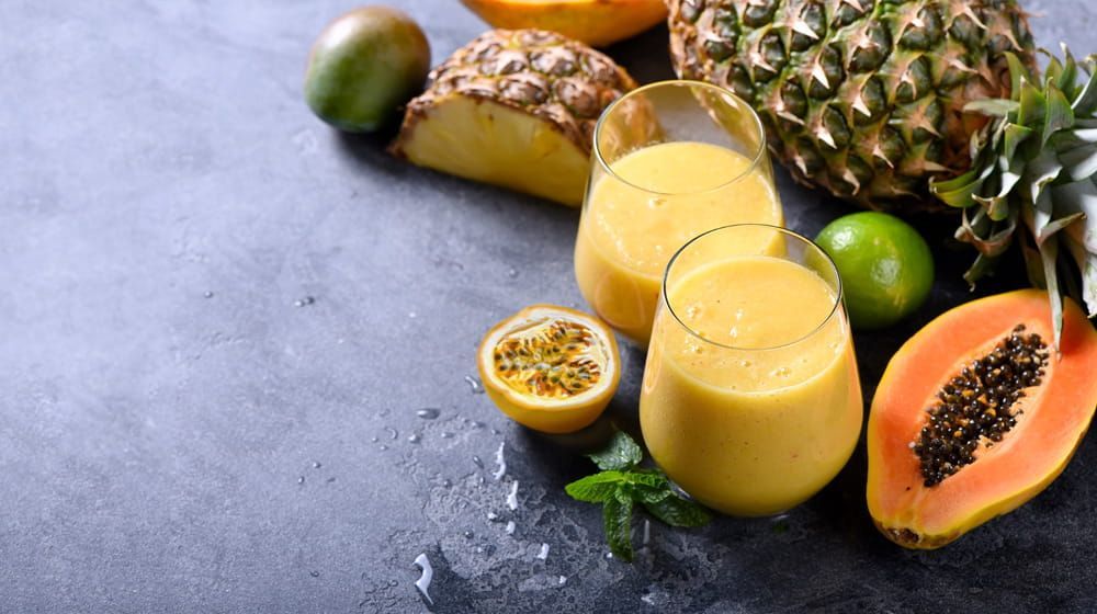 Frutas tropicales y dos batidos amarillos sobre una superficie de piedra oscura, con piña, plátano, lima y maracuyá.