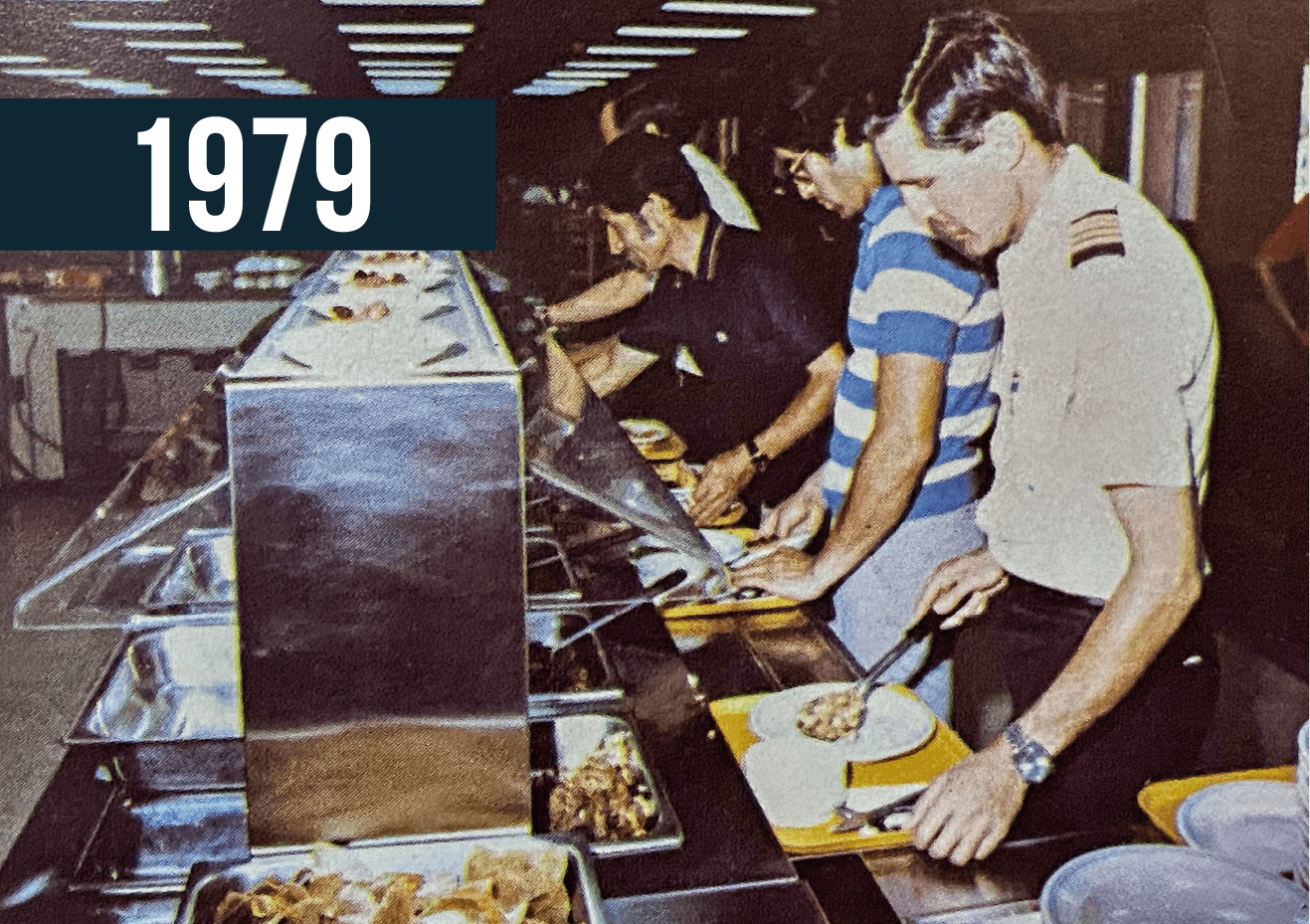 Línea de buffet de 1979 con dos personas sirviéndose la comida bajo luces brillantes del techo.