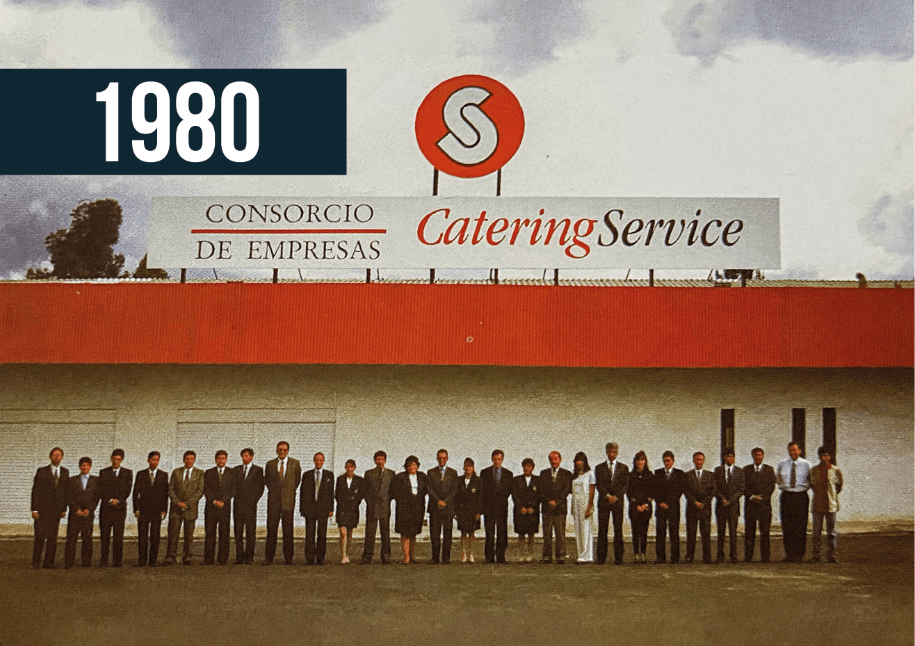 Foto de grupo del servicio de catering de 1980: el personal posa frente a un edificio con techo rojo.