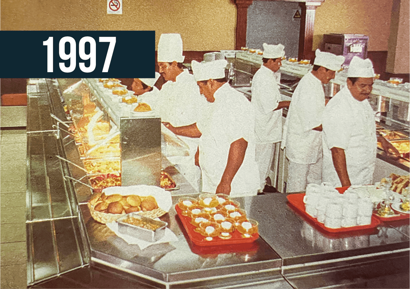 Personal de la cafetería en 1997 sirviendo comida detrás de un mostrador de acero inoxidable.