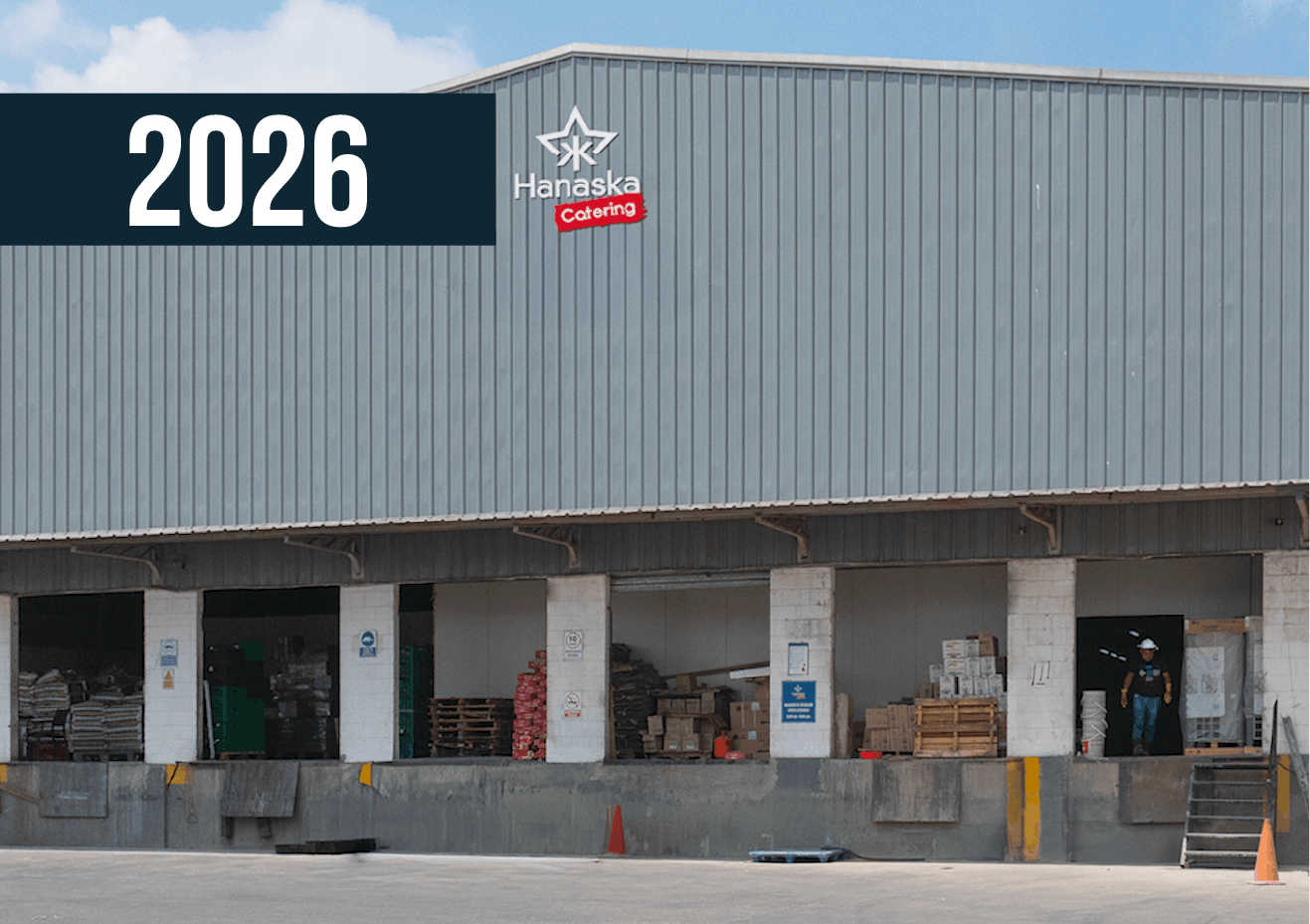 Fachada de tienda con estructura de chapa ondulada gris y muelles de carga, con la inscripción “2026” en la parte superior izquierda.