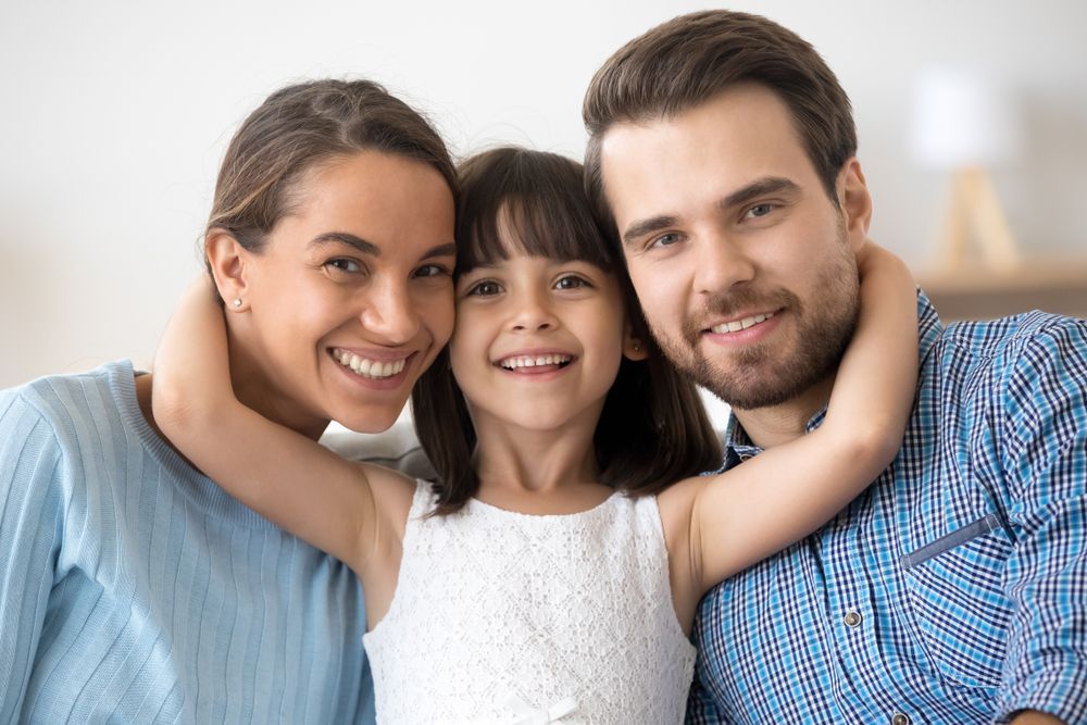 A child smiles while hugging their parents in a bright indoor setting.