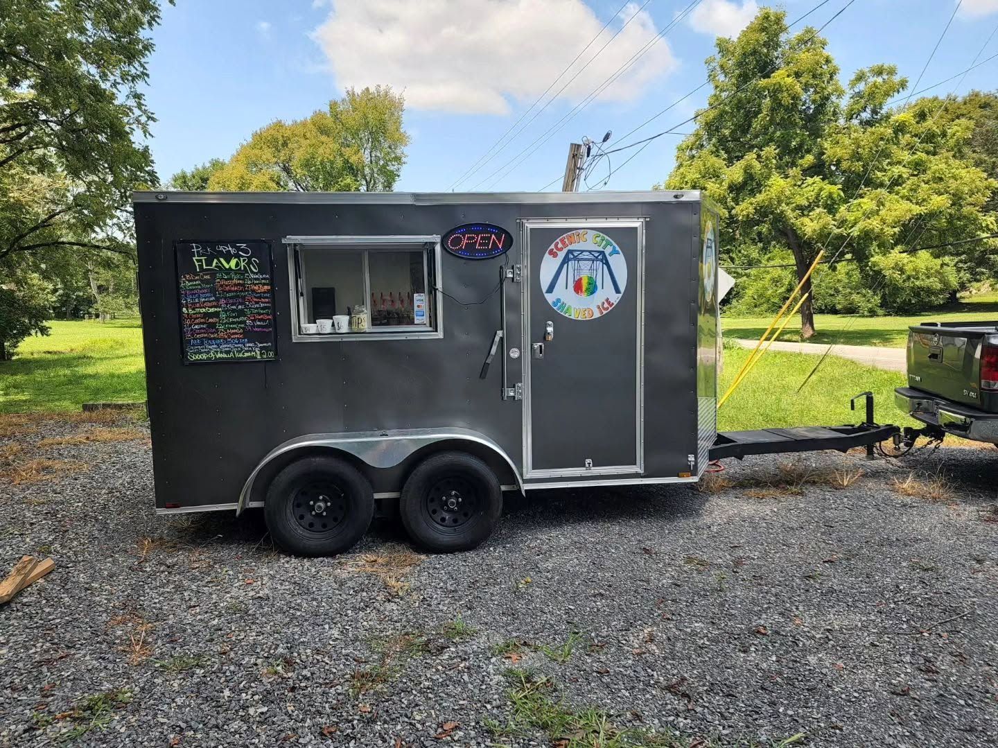 Black food truck trailer with serving window, parked on gravel under trees, with a colorful logo on the door