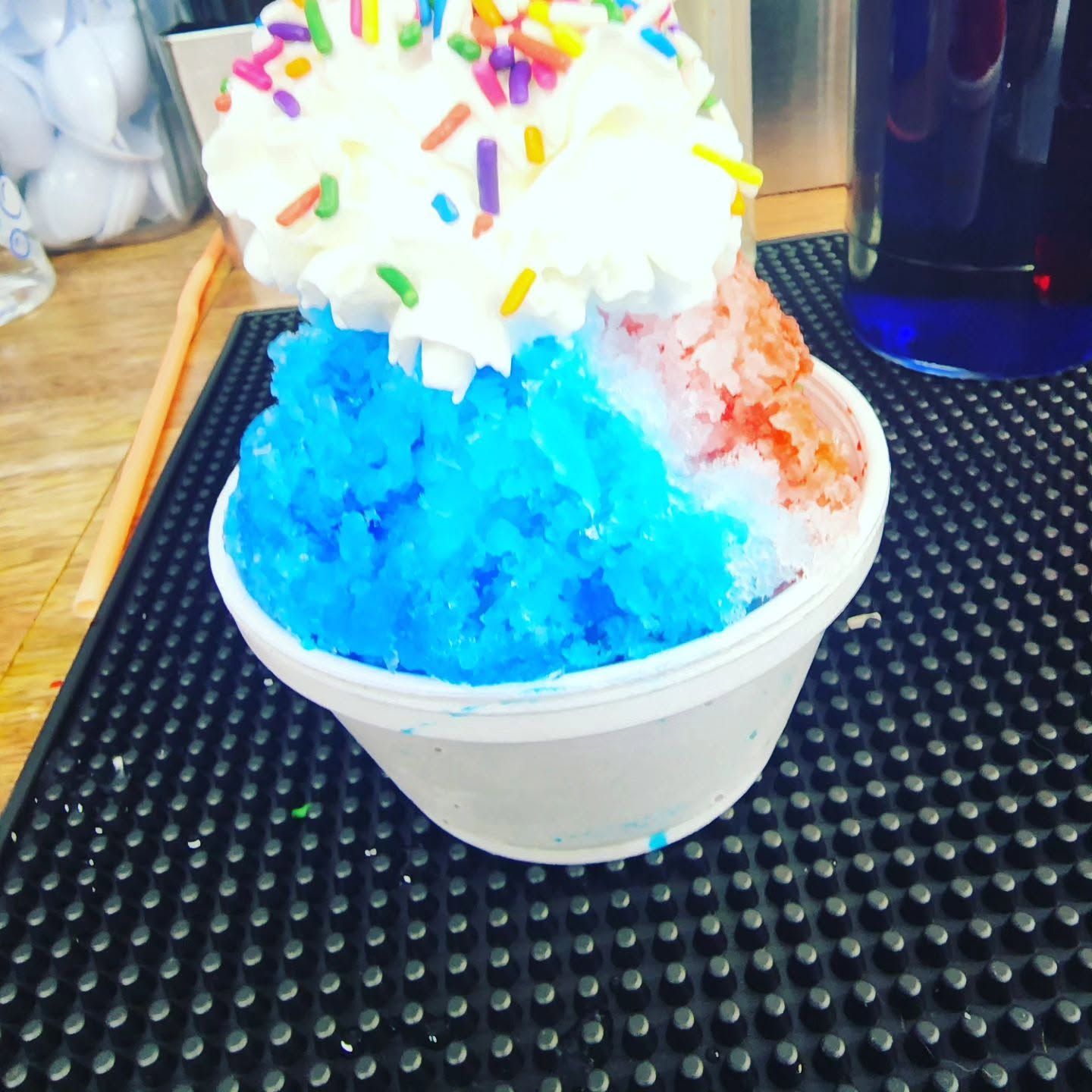 Colorful shaved ice dessert in a white cup with whipped cream and sprinkles on a counter
