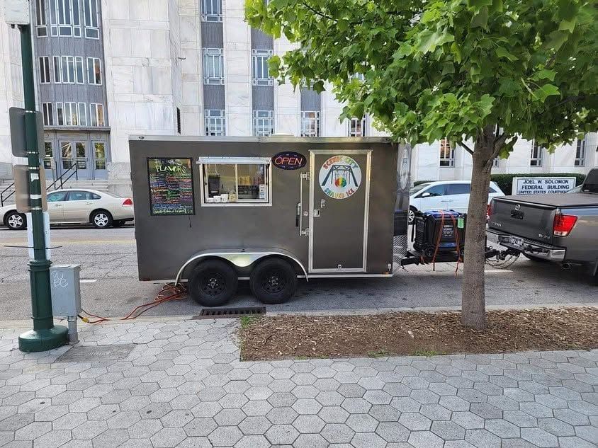 Small gray food trailer parked on a city sidewalk beside trees and apartment buildings.