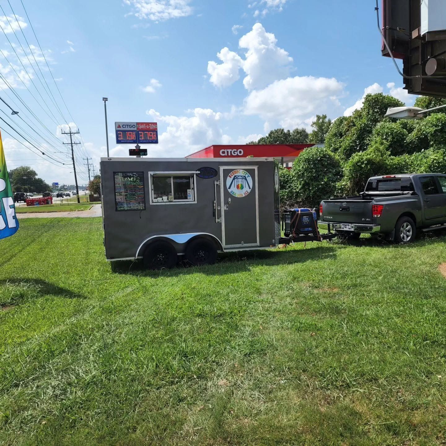 Gray food trailer on a grassy roadside lot beside a parked pickup truck under a bright blue sky.