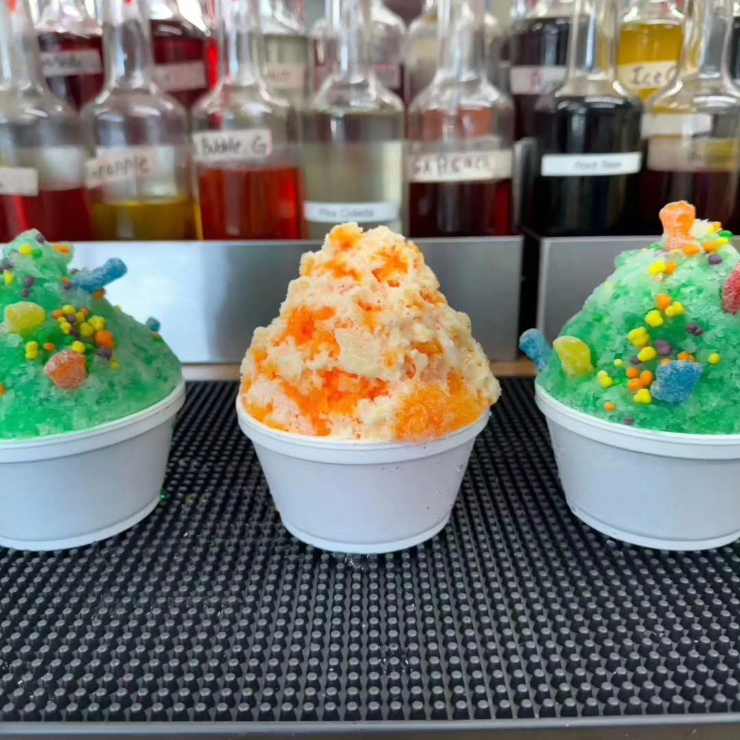 Three colorful shaved-ice desserts in white cups on a bar counter, with bottles blurred behind.