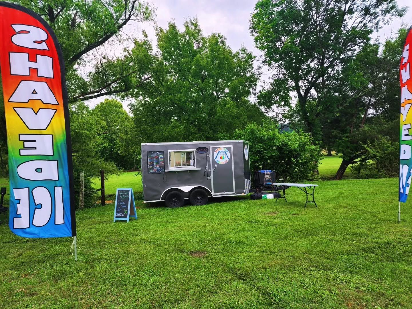 Small black food trailer on grass at a festival, flanked by colorful vertical flags and trees.