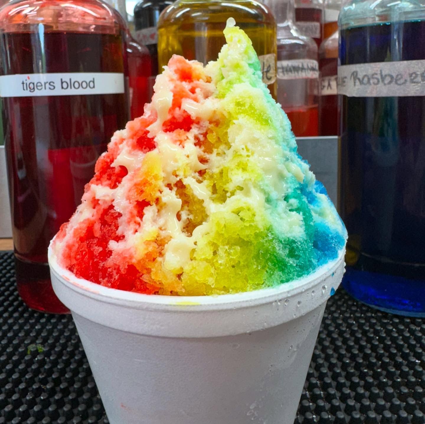 Rainbow shaved ice in a white cup with syrup bottles in the background