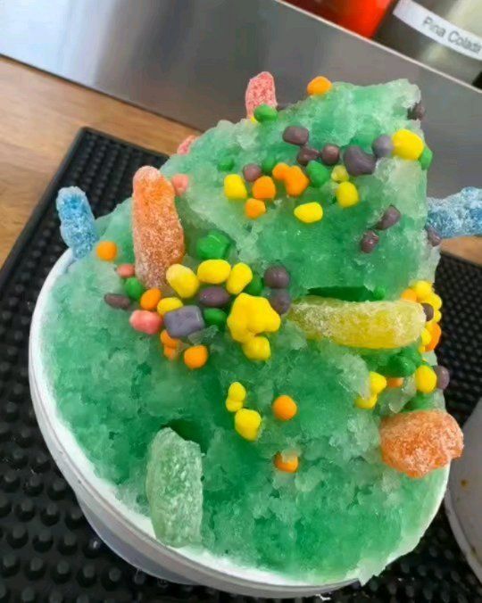 Green shaved ice with colorful sprinkles and gummy candy in a white cup.