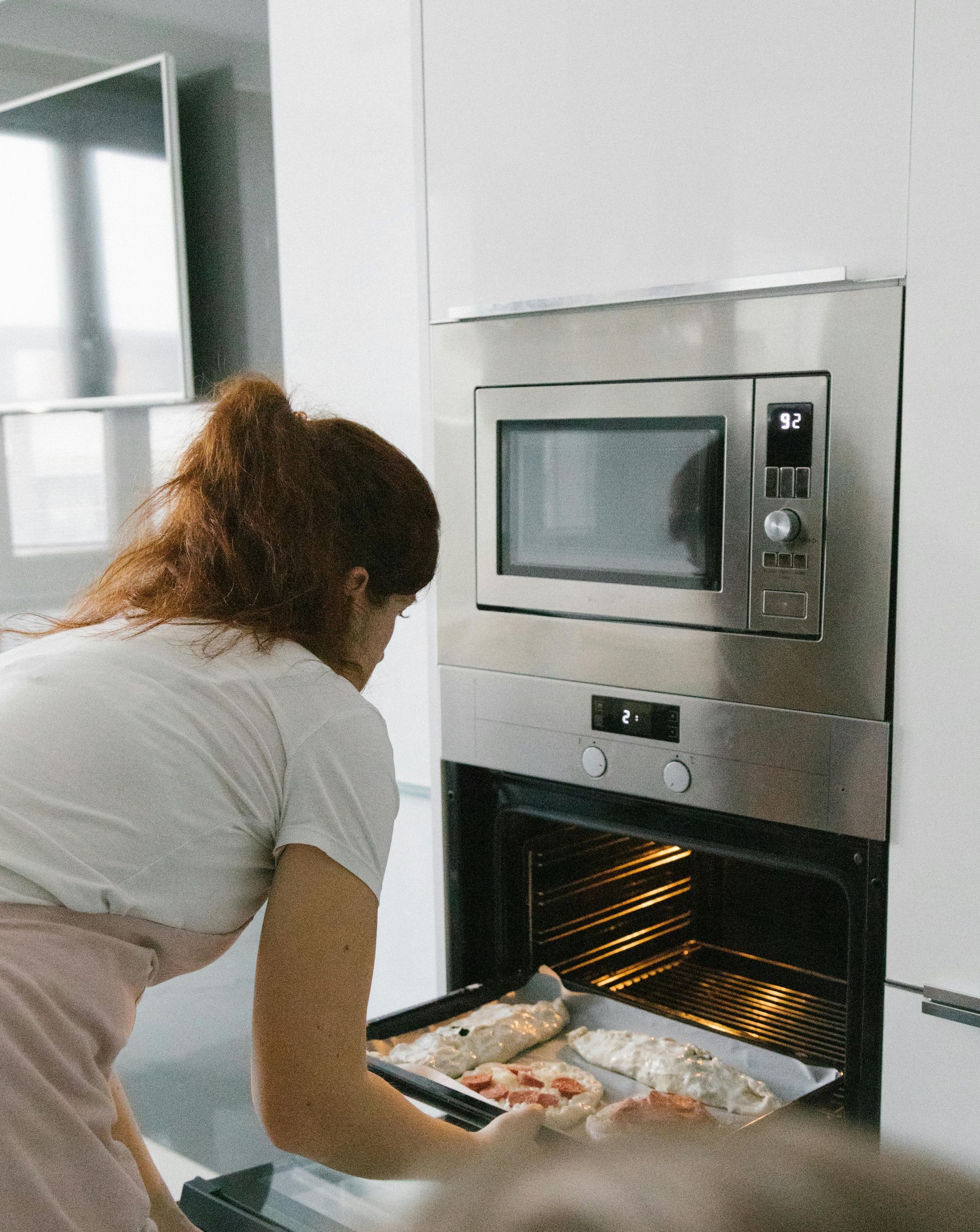 Woman taking food out of an oven in a modern kitchen. She wears a white top and pink apron.