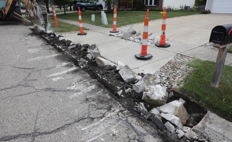 Sidewalk and road construction; concrete and asphalt being broken up, orange cones in place.