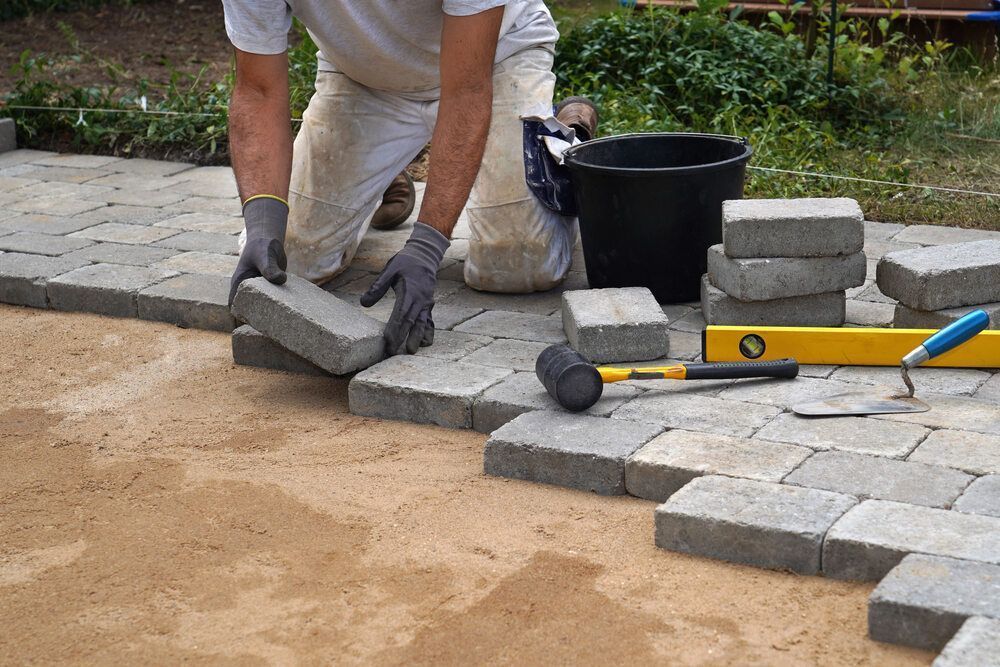 Person laying interlocking brick pavers on a bed of sand outdoors, with tools and supplies nearby.