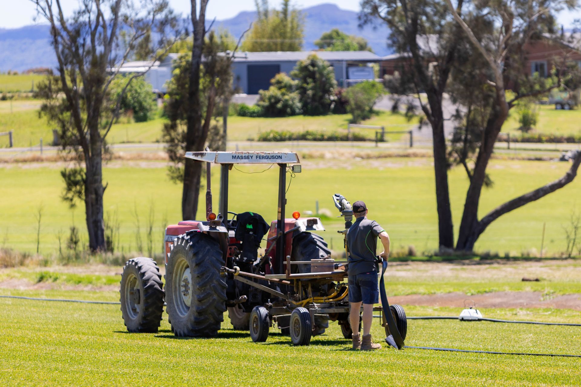 Man with a shovel standing near a tractor on a grassy field; rural landscape in background.