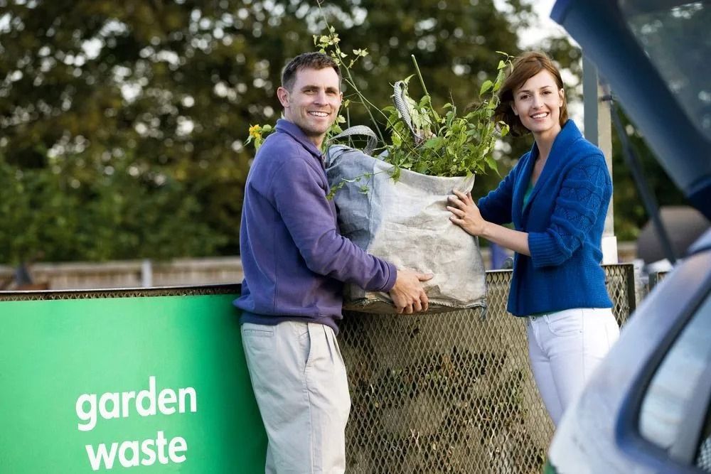 A man and woman smile while depositing garden waste. They stand beside a green bin labeled